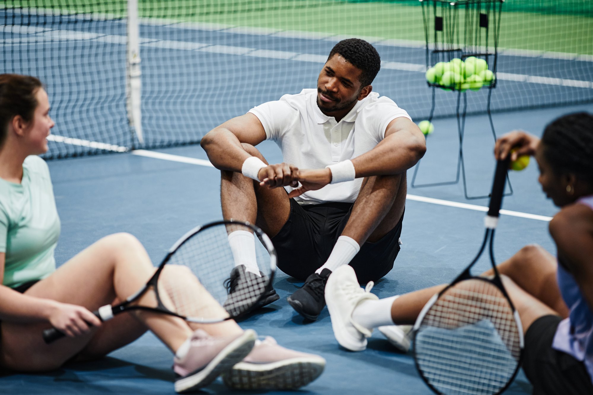 3 adult tennis players sitting on a court with a ball basket in the background